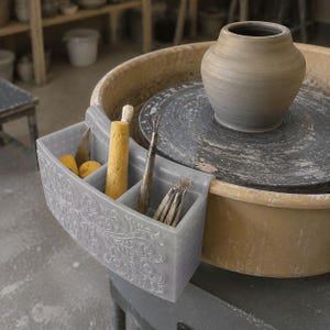 Pode incluir: Um organizador de ferramentas de cerâmica de plástico cinza-claro com padrão floral em relevo fixado a uma roda de oleiro. O organizador contém uma esponja amarela, uma costela de metal e ferramentas de madeira. Um vaso de argila cinza está na roda.