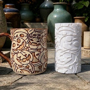 Può includere: Una tazza in ceramica con manico smaltato marrone e un motivo a zucche intagliate, accanto a uno strumento cilindrico bianco con lo stesso design in rilievo. Entrambi gli oggetti presentano un motivo ripetuto di facce di zucca.