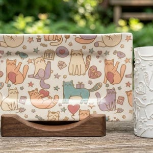 Op de afbeelding: Een rechthoekig keramieken dienblad met een herhalend patroon van kleurrijke katten, hartjes, sterren en cadeautjes, geplaatst op een donkerhouten standaard naast een witte cilindrische deegroller met een reliëf van katten en hartjes.