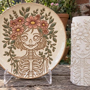 May include: A round ceramic plate and a cylindrical ceramic vase, both featuring a relief design of a skeleton figure adorned with a floral crown and foliage. The plate has a speckled cream finish with pink flowers and green leaves.