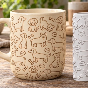 Può includere: Una tazza in ceramica color crema e un rullo cilindrico per texture in argilla bianco, entrambi caratterizzati da un motivo in rilievo di varie razze canine, piccoli ossi e ciotole per cibo. La tazza ha un manico curvo a sinistra.