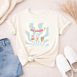 Könnte beinhalten: Ein cremefarbenes Kurzarm-T-Shirt mit einer zentralen Grafik aus hellblauen und blassgelben, wirbelnden Strahlen. Das Design zeigt Illustrationen eines Eis am Stiel, einer Eistüte und eines Eisbechers mit dem Text Let's Go Ice Cream.