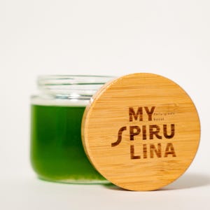 May include: A clear glass jar filled with a vibrant green liquid, accompanied by a round bamboo lid. The lid features the laser-engraved text MY SPIRU LINA and the smaller text daily green boost centered on the wood grain surface.