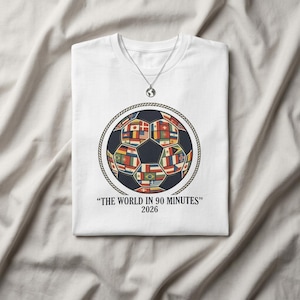 Puede incluir: Camiseta blanca con un diseño de balón de fútbol compuesto por varias banderas nacionales dentro de un marco circular. La camiseta incluye el texto "THE WORLD IN 90 MINUTES" y el año 2026. También se ve un collar plateado con un colgante de globo.