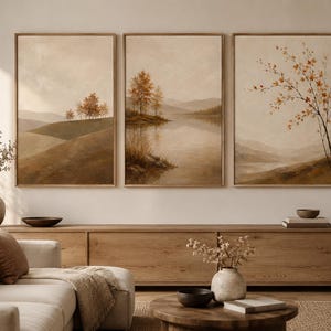 Könnte beinhalten: Ein Set aus drei rechteckigen Wandbildern mit Landschaftsmalereien in gedeckten Erdtönen. Die Kunstwerke zeigen sanfte Hügel, einen ruhigen See und vereinzelte Bäume mit herbstlich gefärbtem Laub, gerahmt in hellem Holz.