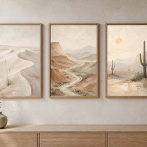 May include: A set of three framed wall art prints featuring desert landscapes. The first print shows sand dunes, the second depicts a winding path through rocky canyons, and the third displays a desert scene with cacti and a sun in a pale sky.