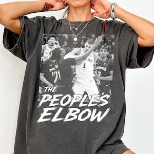 Puede incluir: Camiseta gris oscuro con una imagen en blanco y negro de jugadores de baloncesto. El texto "THE PEOPLE'S ELBOW" está impreso en blanco. La camiseta tiene mangas cortas y un corte holgado.