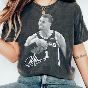Puede incluir: Camiseta gris oscuro con una imagen en blanco y negro de un jugador de baloncesto sosteniendo un balón. El jugador lleva una camiseta con el número 1 y una firma. La camiseta es de estilo vintage.