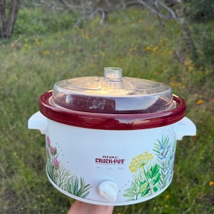 May include: A white Rival Crock-Pot stoneware slow cooker with a maroon rim and a clear glass lid. The exterior features botanical illustrations of herbs and the text Rival Crock-Pot Stoneware Slow Cooker with Off, Low, and High settings.