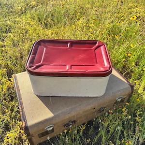 May include: A vintage white and red metal container with a rectangular shape and a matching red lid. The container is placed on top of a tan suitcase with brown trim, set against a backdrop of wildflowers.