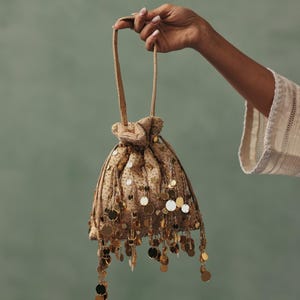 May include: A beige drawstring bag adorned with gold sequins and hanging gold coin details. The bag is held by a person with a light skin tone against a muted green backdrop. The bag's design evokes a vintage aesthetic.