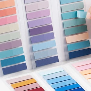 May include: A collection of rectangular adhesive page markers arranged in vertical strips on clear plastic backing. The markers feature a diverse palette including shades of peach, pink, lavender, purple, blue, teal, and navy, with a matte finish.