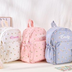 May include: Three backpacks in white, pink, and light blue, each featuring a small floral pattern. The white bag is personalized with the name Raven, the pink bag with Sophie, and the light blue bag with Emily in white or pink script text.