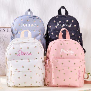 May include: Four backpacks in light blue, black, white, and pink, each featuring a small floral pattern. The backpacks are personalized with the white or pink embroidered names Jennie, Kristy, Stella, and Melody on the front zippered pocket area.