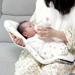 May include: A baby is held in a white, cushioned baby feeding support with a gray heart pattern. The baby is being fed from a clear bottle with a white top. The baby is wearing a white onesie.