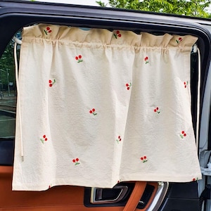 May include: A cream-colored car window curtain featuring a repeating embroidered pattern of small red cherries with green stems. The fabric is gathered at the top with a ruffled header and is suspended by thin white cords on each side of the window.