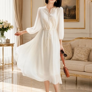 May include: A white midi dress featuring long sleeves, a split neckline, and a central panel with intricate embroidery. The waist includes a drawstring tie with pearl-like accents, and the skirt flows into a loose, lightweight hemline.