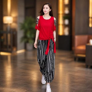Puede incluir: Una blusa roja de manga corta con aberturas en los hombros y tiras negras y blancas con letras, combinada con pantalones holgados de rayas verticales blancas y negras con puños en el bajo y zapatillas deportivas blancas.