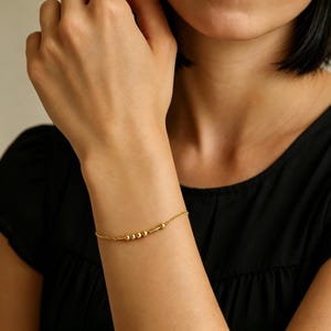 May include: A delicate gold chain bracelet featuring a series of small, polished gold spheres and a short cylindrical bar element. The bracelet is designed with a thin, refined chain that rests smoothly against the skin, showcasing a minimalist aesthetic.