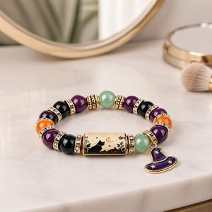 Op de afbeelding: Een kralenarmband met paarse, zwarte, oranje en lichtgroene kralen, afgewisseld met goudkleurige tussenstukjes en strass-ringen. Een centrale rechthoekige goudkleurige kraal toont een zwarte kat en een maansikkel met een paarse heksenhoed.