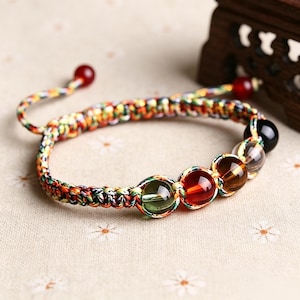 May include: A braided cord bracelet featuring a series of smooth, round, multi-colored translucent beads in shades of green, red, brown, clear, and black. The adjustable band is woven with multicolored threads and finished with two small red beads.