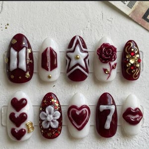 May include: A set of ten artificial nails in various designs. The nails feature a mix of white and burgundy colors, with embellishments like hearts, stars, flowers, and the letter "B". Some nails have gold accents and glitter.