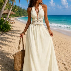 Puede incluir: Un vestido largo de color crema con cuello halter, escote en V profundo, cuerpo adornado con detalles de conchas marinas colgantes y falda fluida. El vestido se combina con un bolso de paja trenzada y un collar con un gran colgante de concha.
