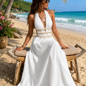 Puede incluir: Un vestido blanco largo hasta el suelo con cuello halter, escote en V profundo, espalda abierta y cintura adornada con detalles de conchas colgantes. El vestido se combina con un collar de conchas a juego y pulseras doradas en ambas mu&ntilde;ecas.