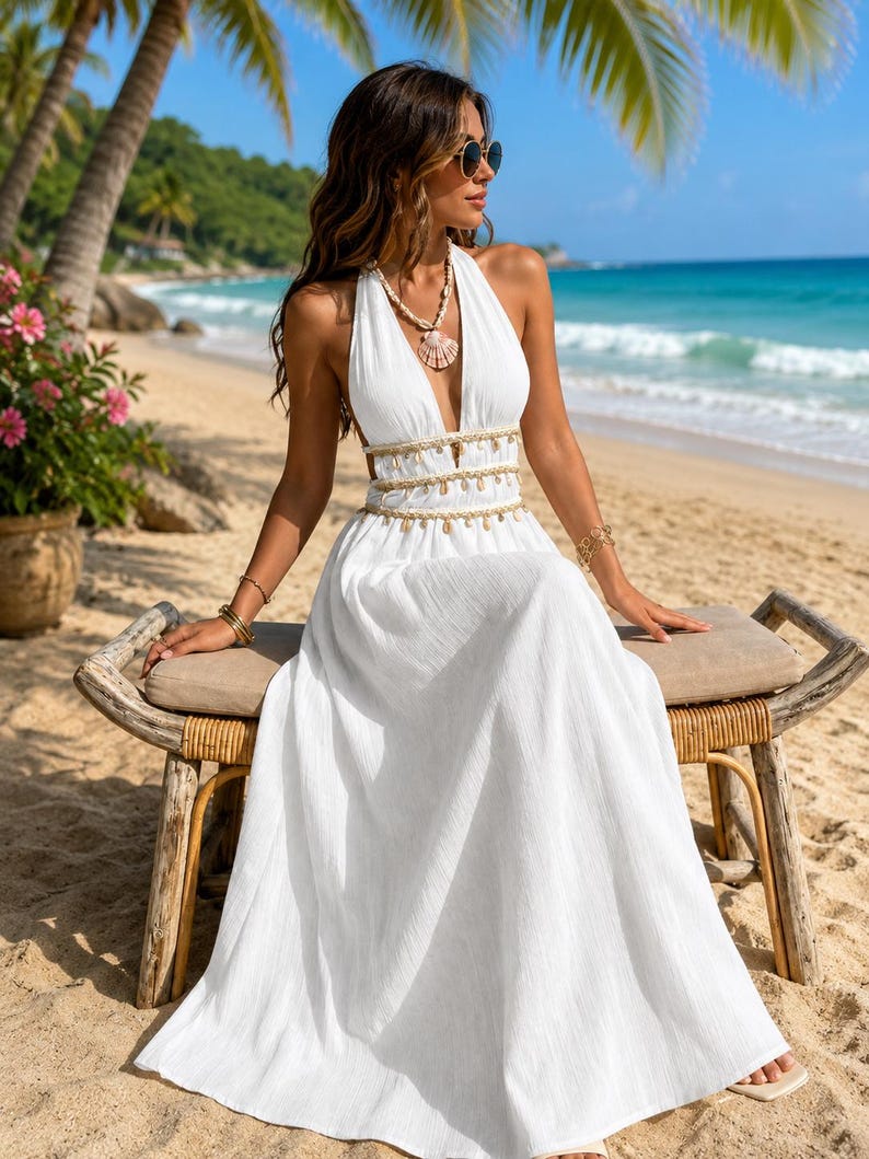 Puede incluir: Un vestido blanco largo hasta el suelo con cuello halter, escote en V profundo, espalda abierta y cintura adornada con detalles de conchas colgantes. El vestido se combina con un collar de conchas a juego y pulseras doradas en ambas mu&ntilde;ecas.
