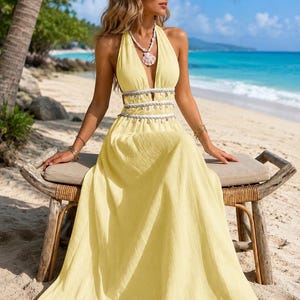 Puede incluir: Un vestido largo de color amarillo p&aacute;lido con un escote en V profundo, un corpi&ntilde;o estilo halter y una cintura con aberturas. La cintura est&aacute; acentuada con dos filas horizontales de flecos de cuentas blancas y plateadas sobre la falda.