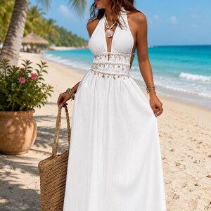 Puede incluir: Un vestido largo blanco tipo halter con escote en V profundo, detalle de cintura con cuentas y adornos de conchas colgantes, y una falda fluida. El vestido se combina con un bolso de paja tejido y un collar de conchas a juego.