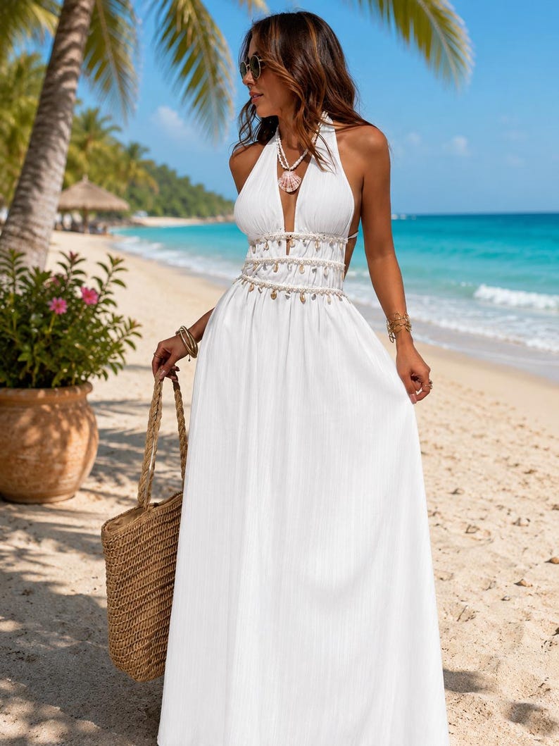 Puede incluir: Un vestido largo blanco tipo halter con escote en V profundo, detalle de cintura con cuentas y adornos de conchas colgantes, y una falda fluida. El vestido se combina con un bolso de paja tejido y un collar de conchas a juego.