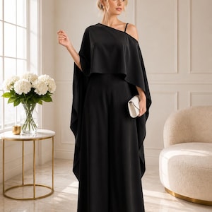 May include: A solid black jumpsuit featuring a wide-leg silhouette and an asymmetrical cape-style overlay that drapes over the shoulders. The garment includes a single thin shoulder strap and is paired with a small, rectangular white clutch bag.