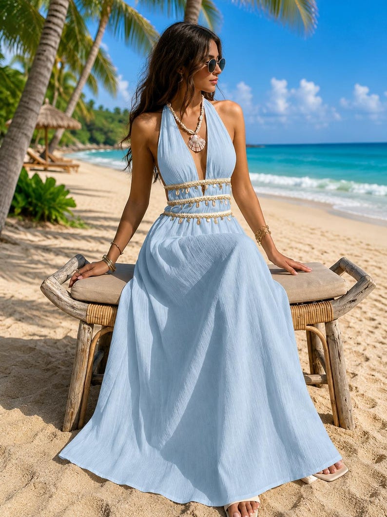 Puede incluir: Un vestido largo de color azul claro con un escote en V profundo, cuello halter y una cintura decorada con adornos dorados y dijes de conchas marinas colgantes. El vestido tiene una falda fluida hasta el suelo con un acabado de tela texturizada.