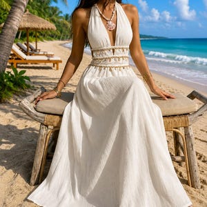 Puede incluir: Un vestido largo blanco hasta los pies con escote halter profundo, acabado de tela texturizada y una cintura adornada con una hilera de peque&ntilde;as conchas marinas colgantes. El vestido incluye un collar de conchas a juego con un gran colgante.