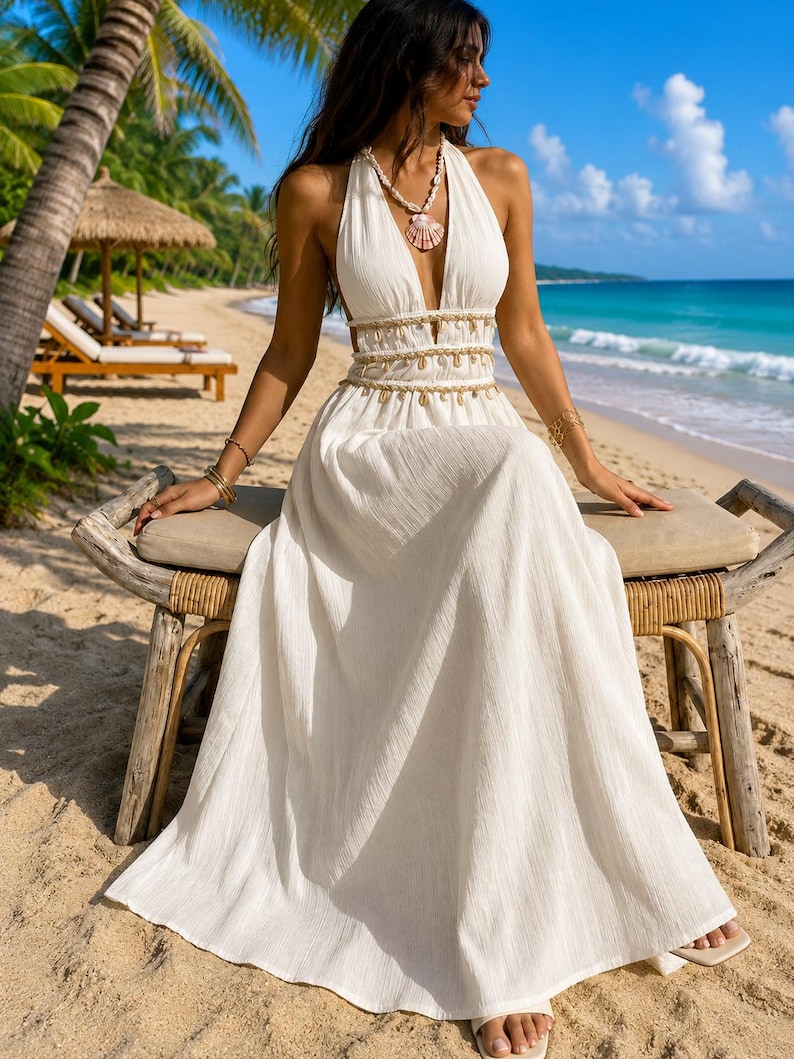 Puede incluir: Un vestido largo blanco hasta los pies con escote halter profundo, acabado de tela texturizada y una cintura adornada con una hilera de peque&ntilde;as conchas marinas colgantes. El vestido incluye un collar de conchas a juego con un gran colgante.