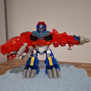 Peut inclure: Une figurine de robot rouge et bleue dotée d'une tête de dinosaure sur le bras droit et d'un accessoire de forage sur le bras gauche. Le personnage porte un casque bleu, des accents jaunes sur les jambes et des pieds en forme de griffes grises.