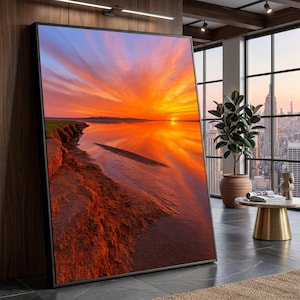 May include: A framed art print showcases a vivid sunset over water. The sky is a fiery mix of orange and yellow, mirrored in the water. The foreground features a reddish-brown shoreline with green grass, creating a dramatic scene.