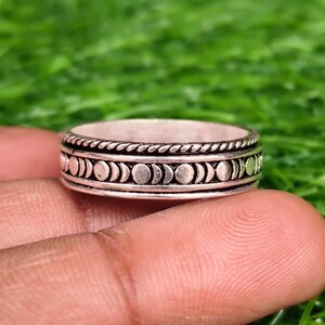 Puede incluir: Un anillo de metal plateado que presenta un patrón repetitivo de fases lunares alrededor de la banda central. El anillo tiene un borde texturizado similar a una cuerda en los bordes superior e inferior, con un acabado pulido que resalta el ciclo lunar grabado.