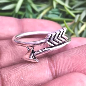 May include: A silver-toned metal ring shaped like an arrow that wraps around the finger. The ring features a detailed fletching design on one end and a pointed arrowhead on the other, with a polished finish and visible dark oxidation in the grooves.