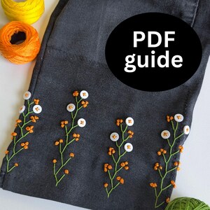 Könnte beinhalten: Dunkelgrauer Stoff mit gesticktem Blumendesign. Die Stickerei zeigt grüne Stiele, orange Knospen und weiße knopfförmige Blüten. Das Bild enthält Spulen mit gelbem, orangefarbenem und grünem Garn. Der Text auf dem Bild lautet "PDF guide" und "Button Flower Embroidery Tutorial."