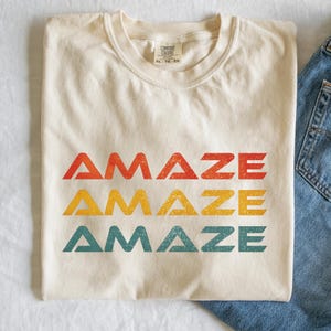 May include: A cream-colored t-shirt featuring the word AMAZE printed three times in a stacked design. The top row is red, the middle row is yellow, and the bottom row is teal. Each word has a distressed, weathered texture and a bold font.