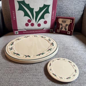 Può includere: Un sottopentola rotondo in ceramica color crema e un sottobicchiere rotondo più piccolo abbinato, entrambi con un motivo di agrifoglio verde e rosso lungo il bordo. Dietro si trova una scatola di cartone quadrata con la scritta TRADITIONAL HOLLY e LONGABERGER POTTERY.