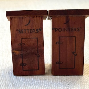 May include: Two brown wooden outhouse-shaped ornaments. The left one features a crescent moon cutout, a door outline, and the text SETTERS. The right one features a crescent moon cutout, a door outline, and the text POINTERS. Both have flat roofs.