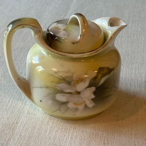 May include: A small ceramic teapot with a lid, featuring a pale yellow and cream gradient background. The body and lid are decorated with hand-painted white flowers and green leaves. It has a curved handle on the left and a small spout on the right.