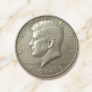 Puede incluir: Una moneda de medio dólar estadounidense de 1964 de color plateado con el perfil de John F. Kennedy mirando hacia la izquierda. La moneda tiene inscrita la palabra LIBERTY en la parte superior, la frase IN GOD WE TRUST abajo y el año 1964.