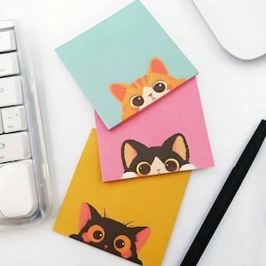 May include: Three square sticky notes featuring cartoon cat illustrations. The top note is light blue with an orange tabby cat, the middle is pink with a black and white cat, and the bottom is yellow with a solid black cat peeking from the bottom.