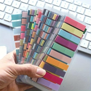 May include: A set of translucent adhesive index tabs arranged in a fan shape. The tabs feature a wide variety of muted and vibrant colors, including shades of pink, blue, green, yellow, orange, and purple, all housed within a clear plastic sleeve.