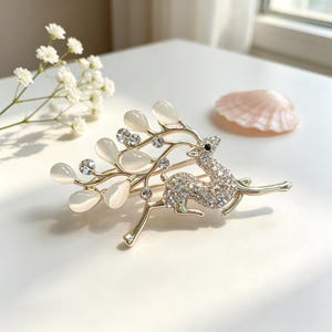 May include: A gold-toned metal brooch shaped like a leaping deer with a body covered in clear rhinestones. The deer's antlers extend into branches adorned with smooth, translucent white oval stones and small clear rhinestones throughout the design.