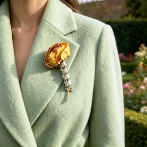 May include: A yellow rose brooch with a stem wrapped in clear, sparkling stones is pinned to a pale green coat. The brooch is a floral accessory, adding a touch of elegance. A garden is visible in the background.